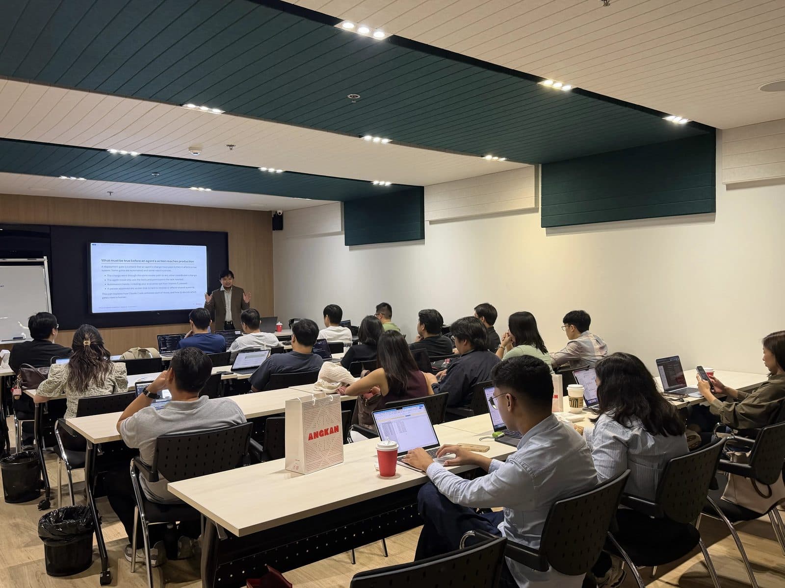A Claude Certified Architect presenting to a classroom of D.W. Morgan engineers working on laptops, with a slide on what must be true before an agent's action reaches production